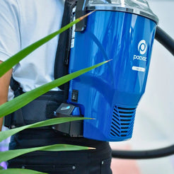 A close-up of a person wearing a blue backpack-style vacuum cleaner with a black harness. The vacuum body is glossy blue with a visible Pacvac logo, and green plant leaves partially frame the shot.