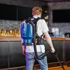 A man seen from behind wearing a blue and black backpack vacuum cleaner in a bar or restaurant setting.