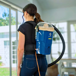 A woman viewed from behind wearing a blue backpack-style vacuum cleaner with a black hose and a clear dust bin, standing indoors near large windows.