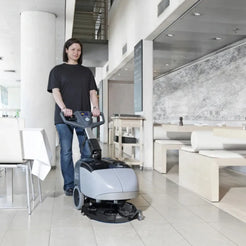 A person in a black t-shirt and jeans stands in a modern indoor lobby, holding the handle of a gray and black commercial floor buffer machine.