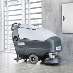 A Nilfisk drive-cleaner vacuum truck for industrial floor cleaning, shown in a modern indoor setting with tiled floors and a concrete wall.