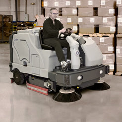 A man wearing safety glasses and a dark uniform sits in the driver's seat of a gray and black OshKosh industrial sweep/driver vehicle, maneuvering it inside a warehouse with stacks of cardboard boxes in the background.