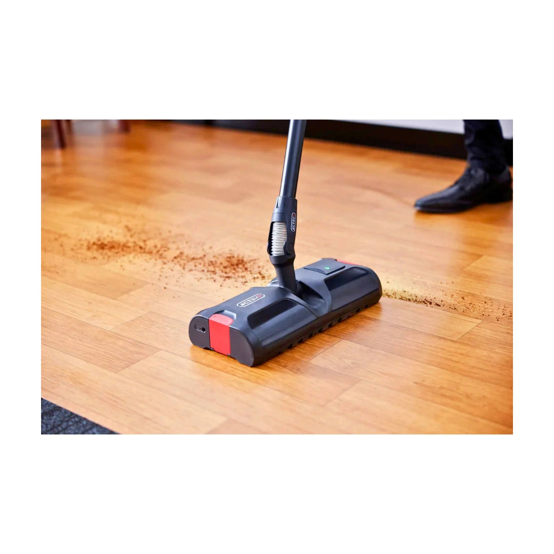 A floor vacuum cleaner head in use on a polished wood floor, lifting up dust and debris, with a person's foot visible in the background.