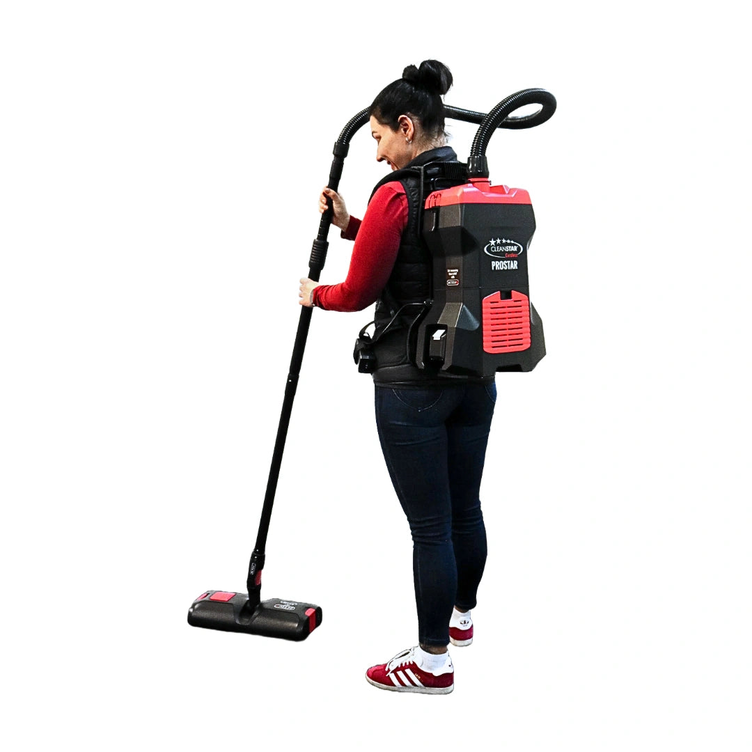 A woman wearing a red long-sleeve shirt, dark jeans, and red-and-white sneakers is shown from the side/back holding a backpack-style cordless vacuum cleaner. The vacuum has a black body with red panels and visible branding that reads 'PROUD' and 'BREATHE BREATHE'.
