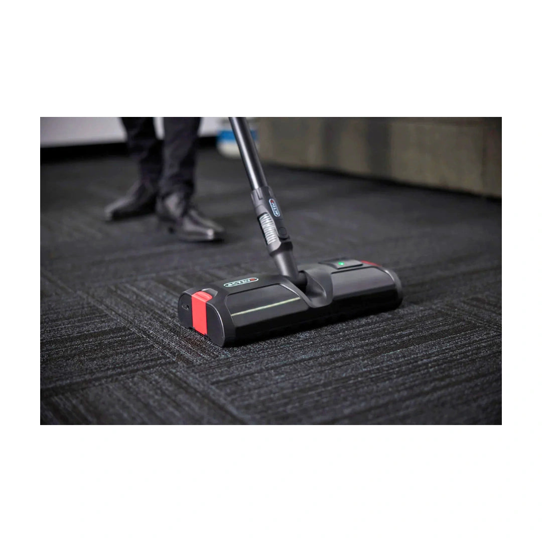 A black and red carpet cleaner head with a black handle is shown cleaning a dark gray textured carpet, with a person's legs and shoes visible in the background.