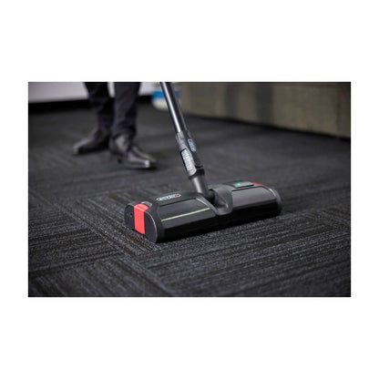 A black and red carpet cleaner head with a black handle is shown cleaning a dark gray textured carpet, with a person's legs and shoes visible in the background.