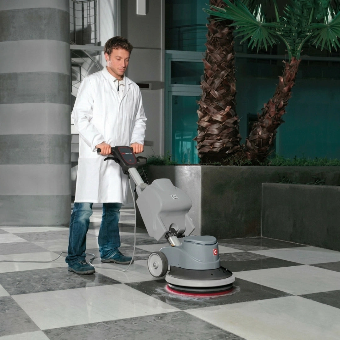 A man in a white lab coat operating a gray floor buffer machine on a polished black-and-white checkered tile floor in a modern indoor setting with palm trees.