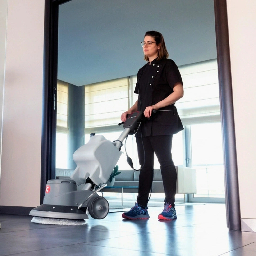 A woman in a black uniform stands in a modern office lobby using a gray and white floor buffer machine.