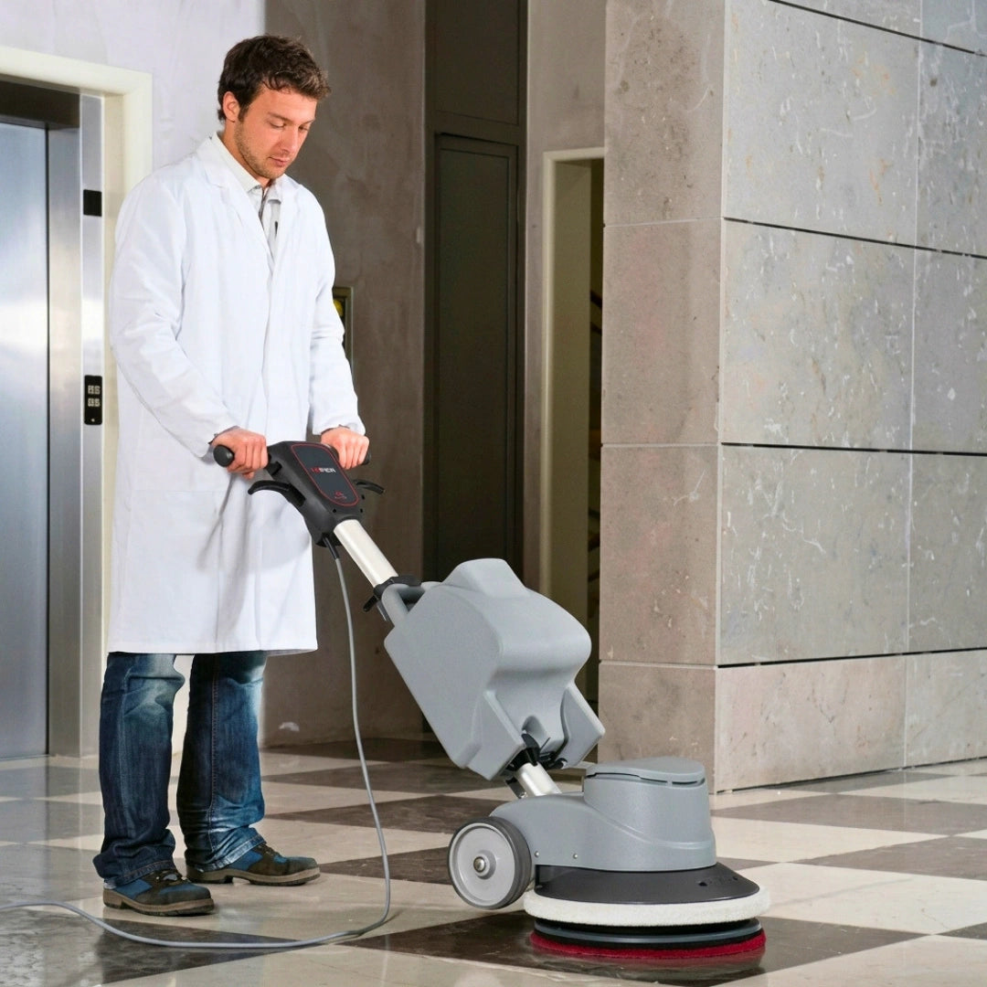 A man in a white lab coat operating a gray and black floor buffer machine in a tiled indoor hallway.