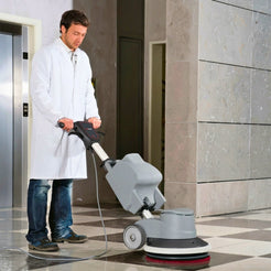A man in a white lab coat operating a gray and black floor buffer machine in a tiled indoor hallway.