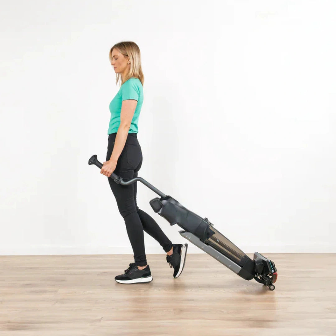 A woman in a teal t-shirt and black leggings walks while pulling a compact black and gray exercise machine with a handlebar and wheeled base on a light wood floor against a white wall.