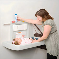 A woman changes a baby's diaper on a wall-mounted changing table. The changing table has a light gray seat and a white storage cabinet with drawers and shelves.