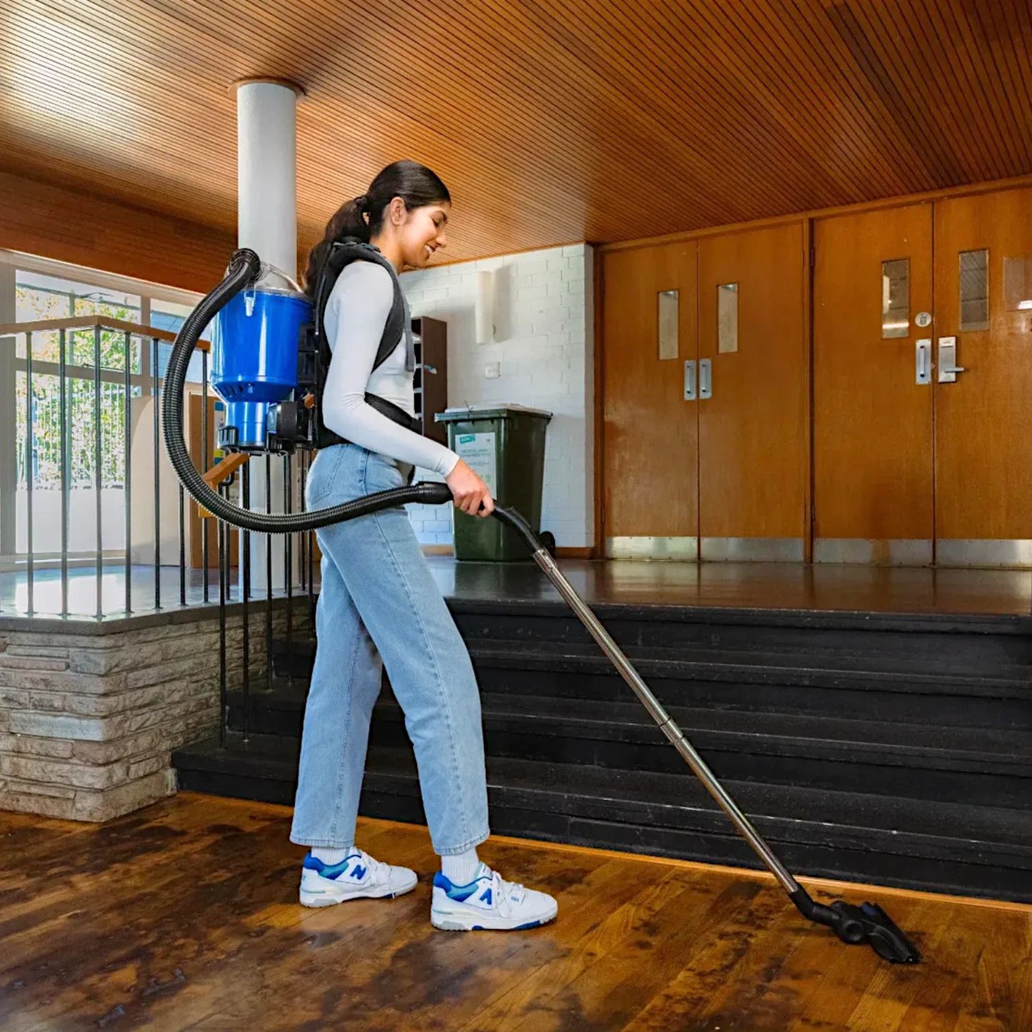 A woman wearing a blue backpack vacuum cleaner is cleaning a wooden floor inside a building with wooden doors and stairs.