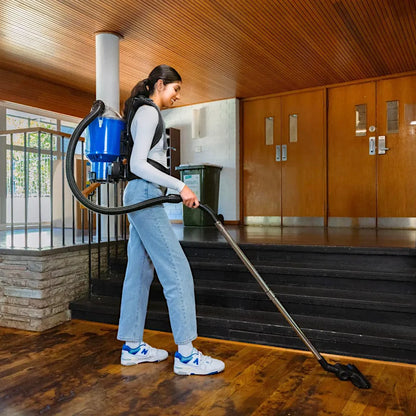 A woman wearing a blue backpack vacuum cleaner is cleaning a wooden floor inside a building with wooden doors and stairs.