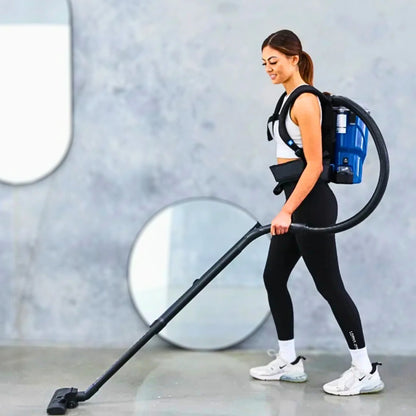 A woman wearing a black and blue backpack vacuum cleaner walks across a modern gray room, holding the vacuum wand. She is dressed in a white sports bra, black leggings, and white sneakers.