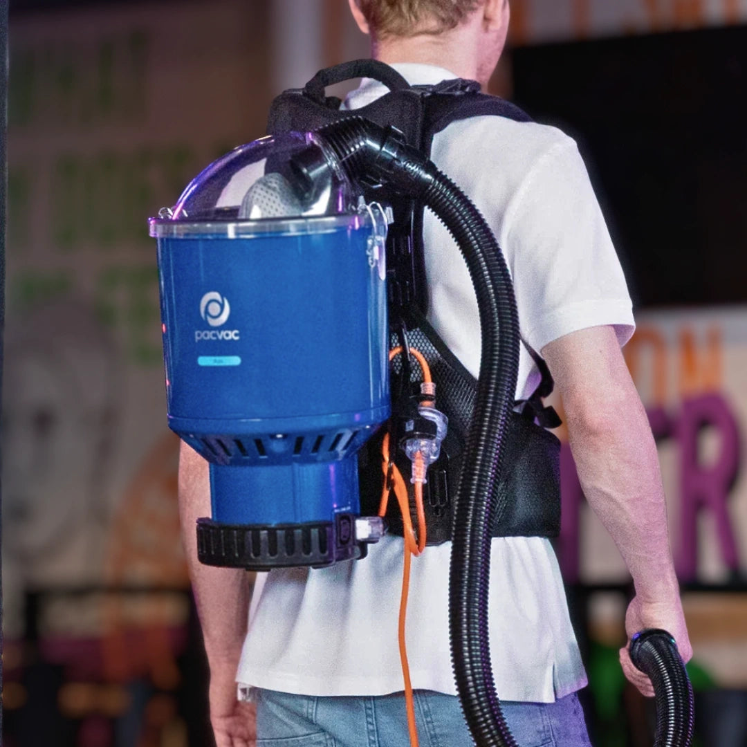 A man viewed from behind wearing a blue and black backpack-style vacuum cleaner with a clear top dome and attached hose.