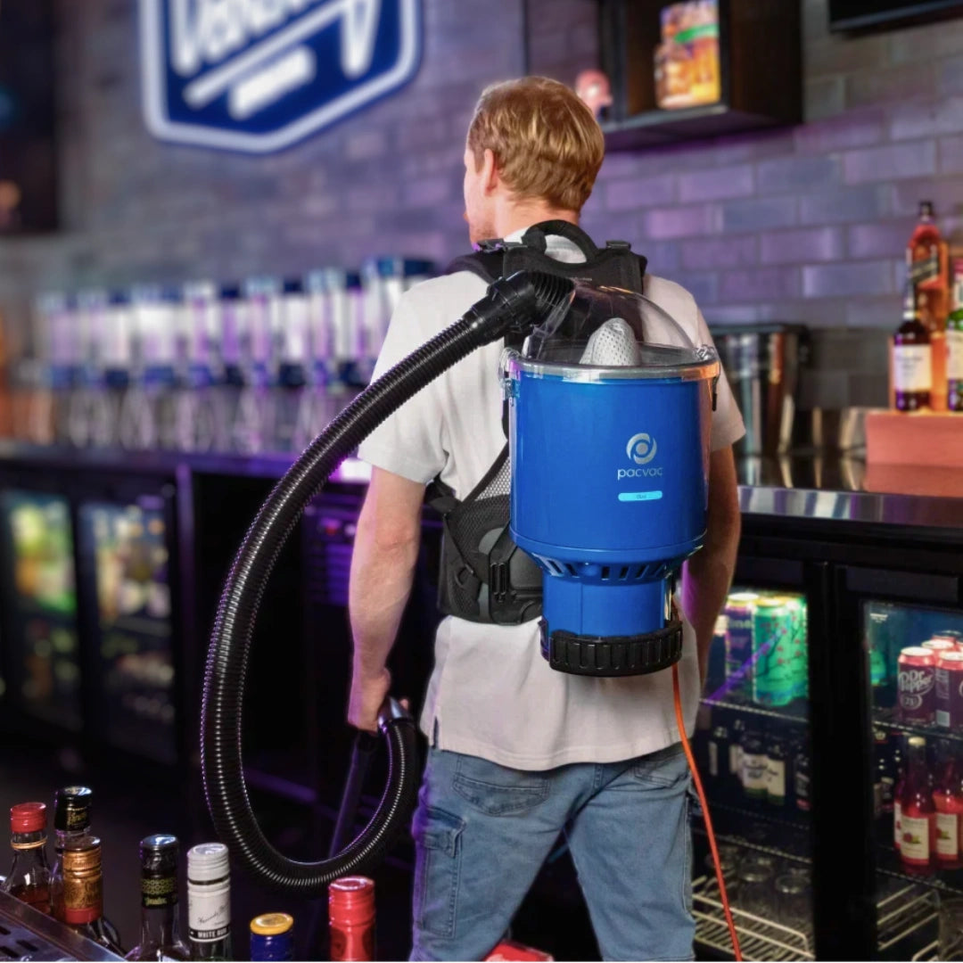 A man standing at a bar, viewed from behind, wearing a blue and black portable back vacuum cleaner with a flexible hose. The bar interior has shelves of bottles and a brick wall in the background.