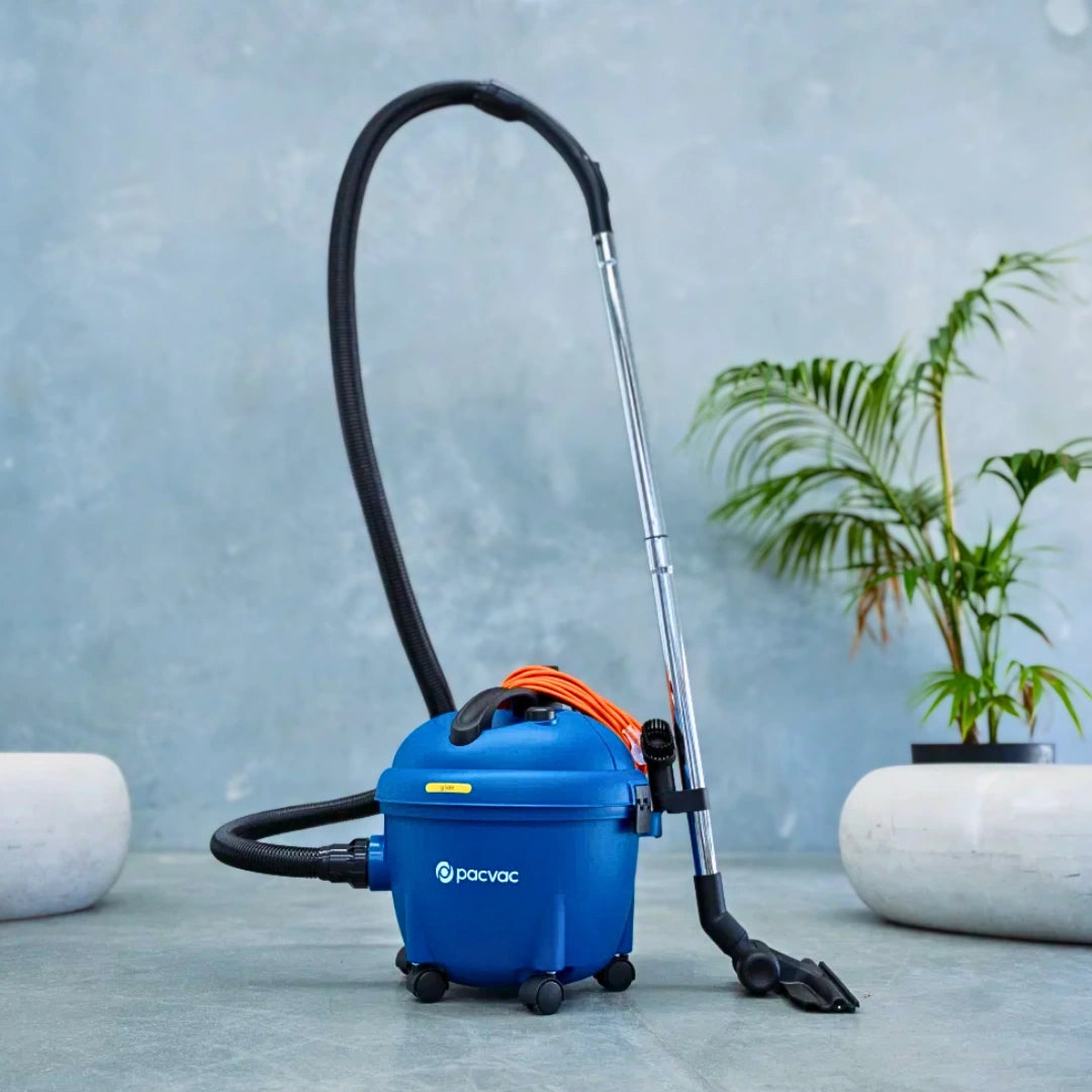 A blue Parvoc wet vacuum cleaner with a black hose, silver wand, and orange cord, set on a gray stone floor in front of a textured blue-gray wall with a potted plant in the background.
