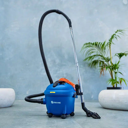 A blue Parvoc wet vacuum cleaner with a black hose, silver wand, and orange cord, set on a gray stone floor in front of a textured blue-gray wall with a potted plant in the background.