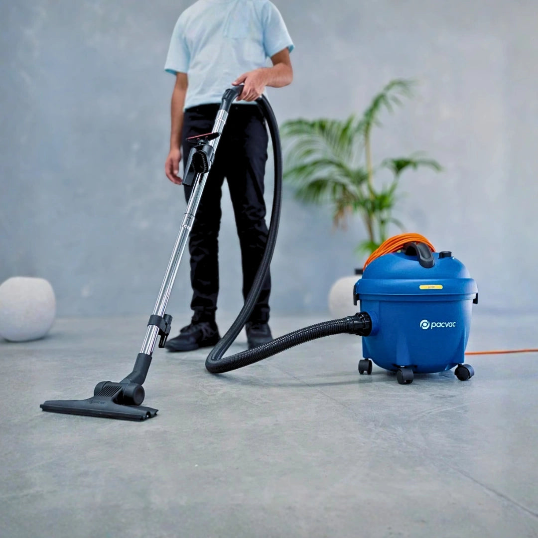 A person using a blue and black vacuum cleaner with an orange handle accent in a modern, minimalist room with a gray concrete floor and wall.