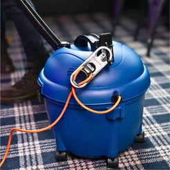 A blue wet/dry vacuum cleaner with an orange cord and a silver-and-black hook-and-loop strap, shown on a plaid carpet with part of a person's leg and foot visible in the background.