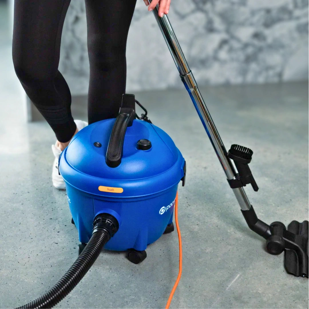 A person in black leggings and white sneakers uses a blue wet/dry vacuum cleaner with a long black wand and hose on a gray concrete floor.
