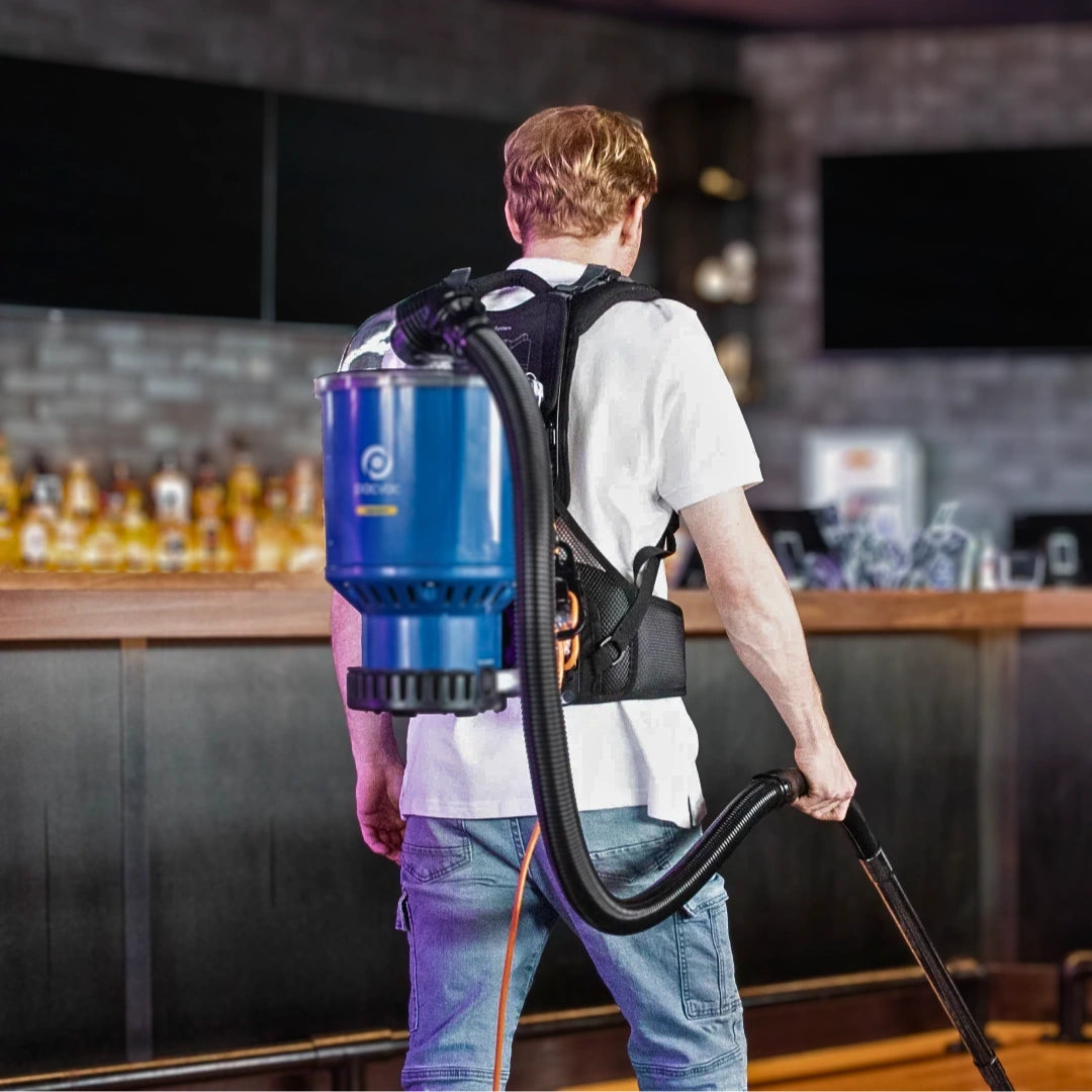 A man seen from behind wearing a blue and black backpack vacuum cleaner in a bar or restaurant setting.