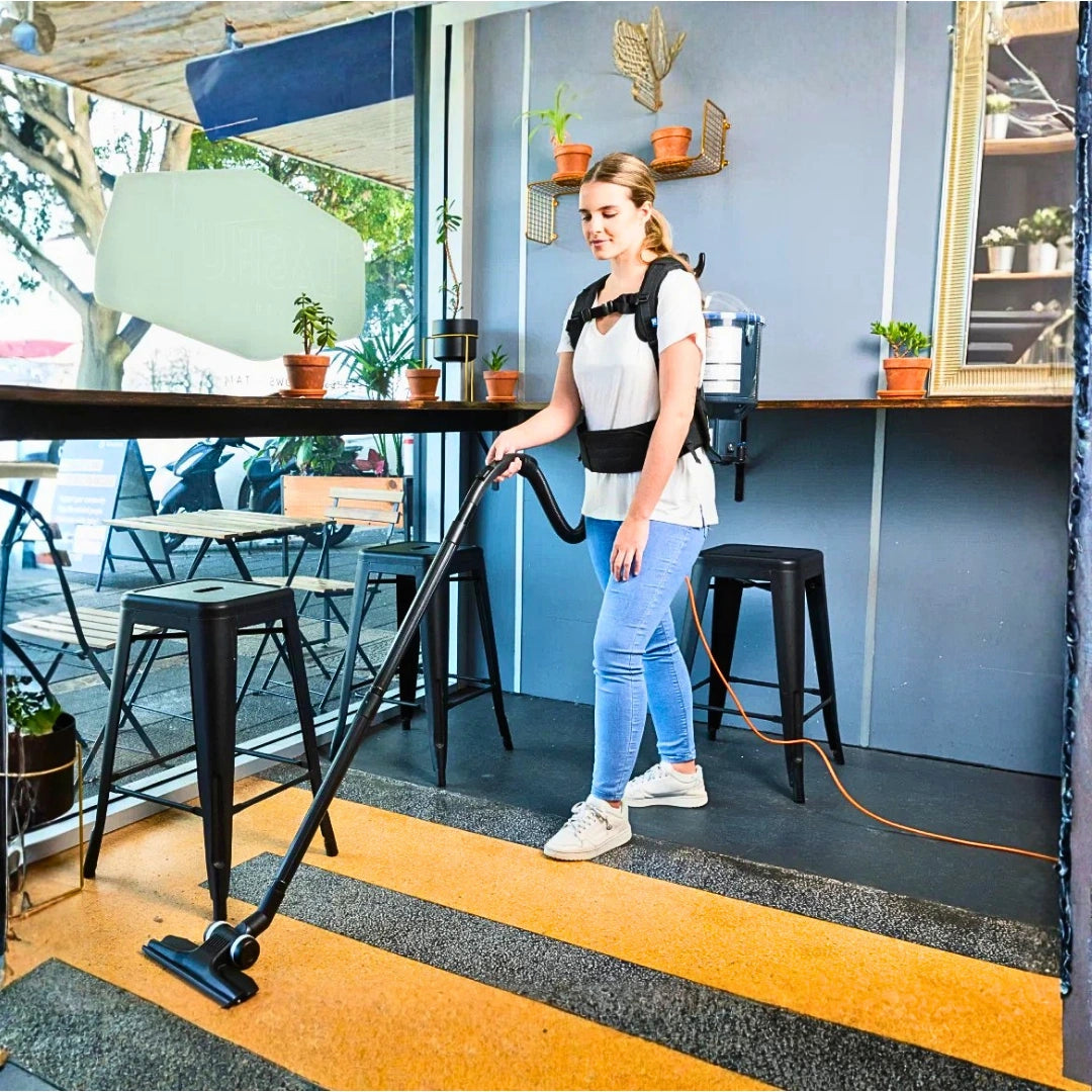 A woman wearing a black and gray backpack vacuum cleaner is cleaning a striped yellow and gray carpet in a modern cafe or bar setting.