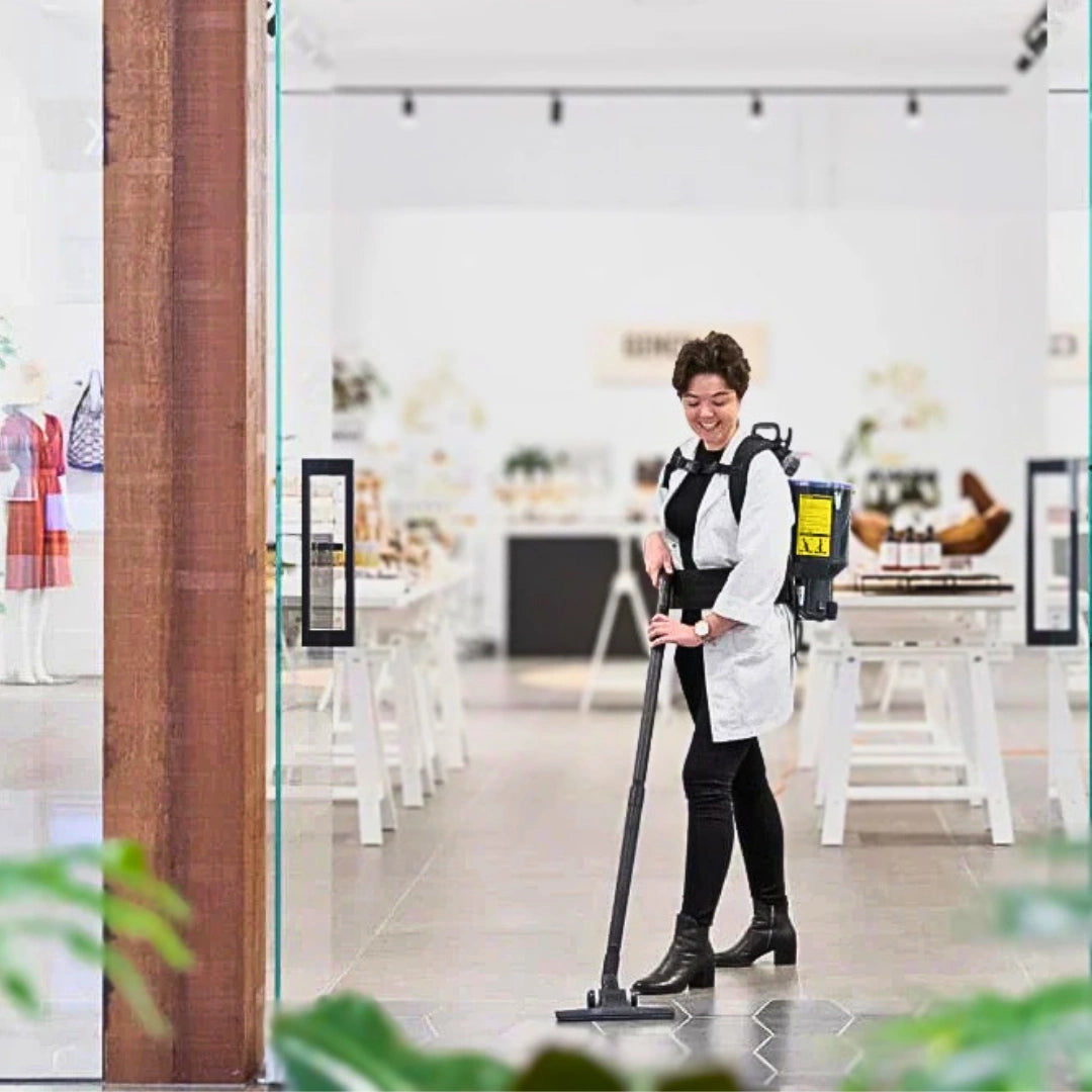 A woman in a white lab coat and black pants is shown using a backpack-style floor cleaner in a bright, modern retail store interior.