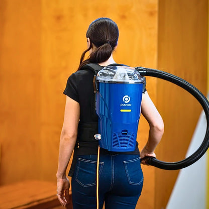 A woman viewed from behind wearing a blue backpack-style vacuum cleaner with a black hose, standing indoors against a warm wood-paneled wall.
