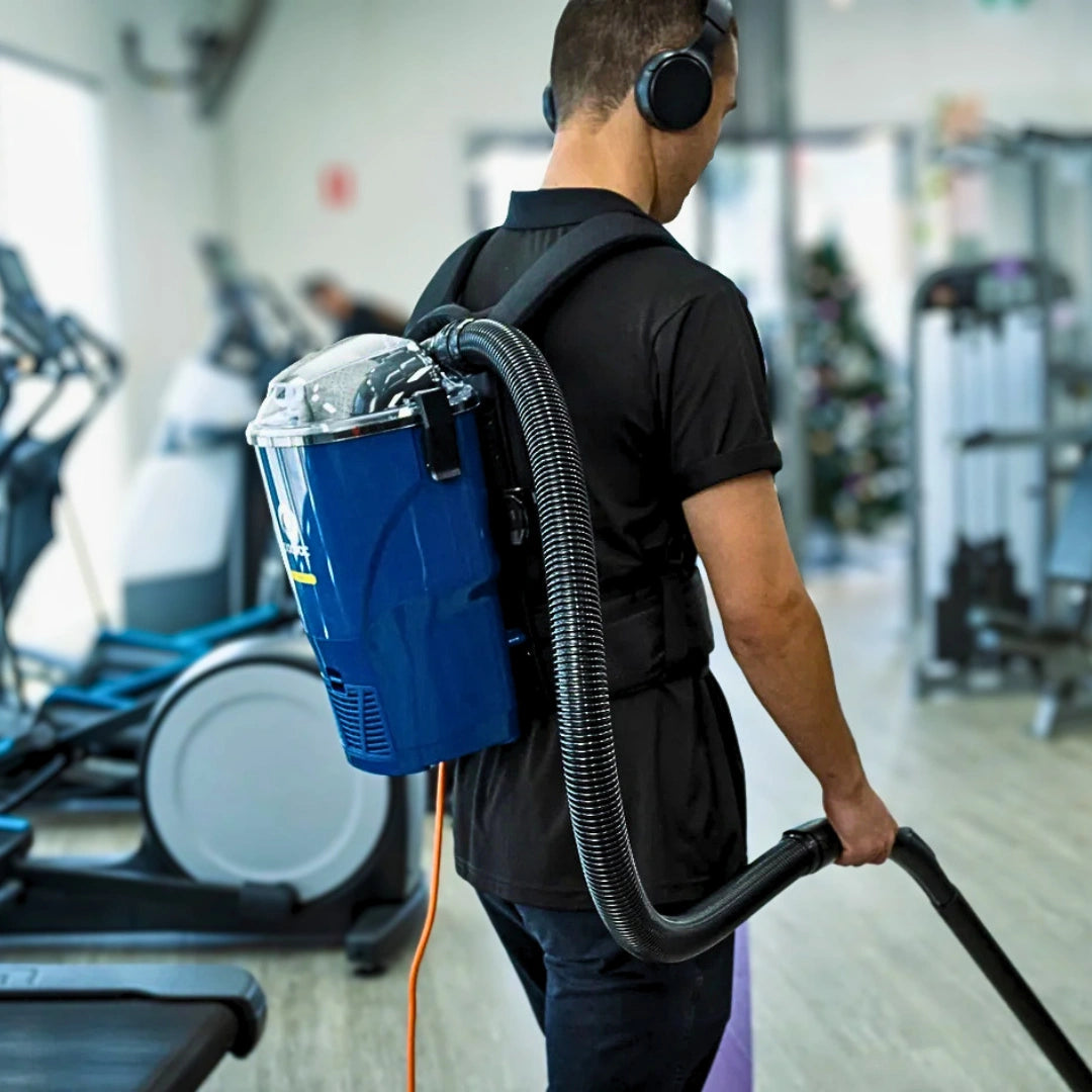 A man in a gym wearing a blue backpack-style floor cleaner with a flexible hose, shown from behind as he operates the device.