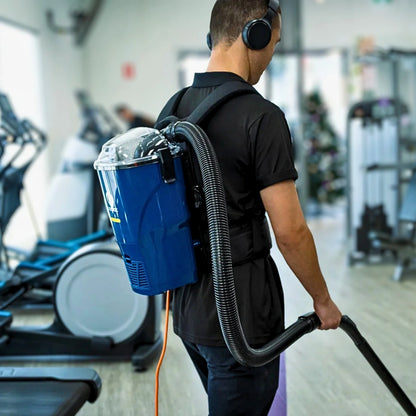 A man in a gym wearing a blue backpack-style floor cleaner with a flexible hose, shown from behind as he operates the device.
