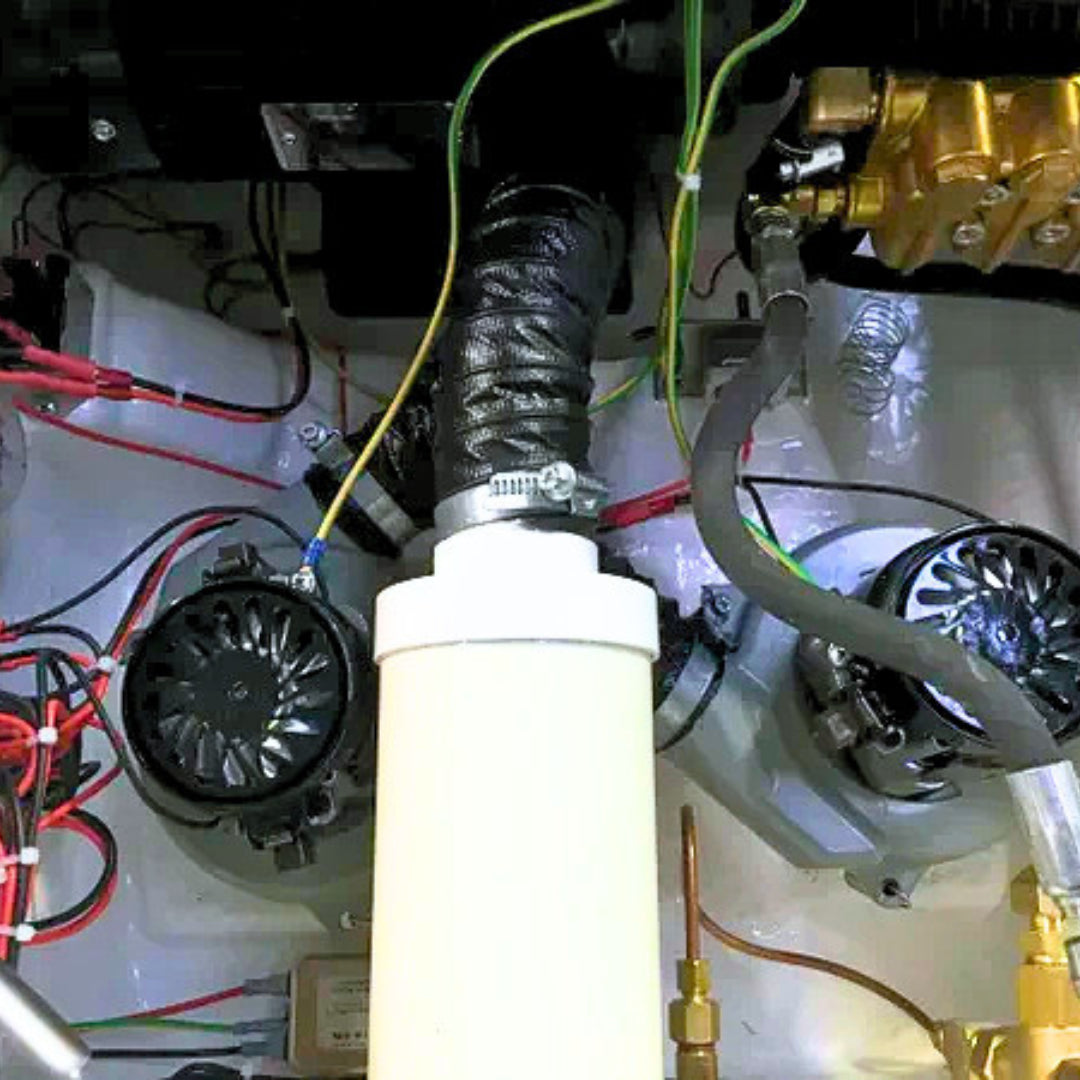 A close-up view of an engine bay component, featuring a prominent white cylindrical filter or canister in the center, connected to a black corrugated hose. Surrounding elements include cooling fans, wiring harnesses, hoses, and metallic engine parts.