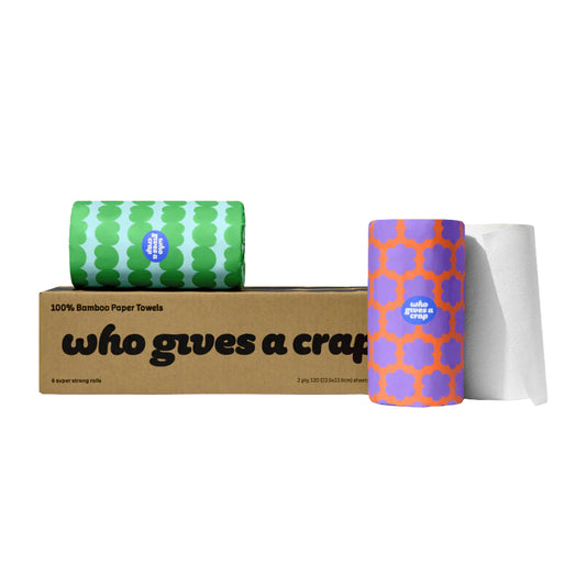 A kraft paper box labeled 'who gives a craft' with two rolls of bamboo paper towels behind and beside it, one roll in green and white pattern and one in purple and orange pattern, with a white towel sheet pulled out on a white background.