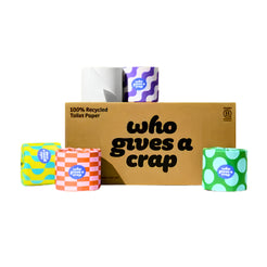 A cardboard box of toilet paper with four colorful rolls arranged in front on a white background. The box reads '100% Recycled Toilet Paper' and 'who gives a crap'.