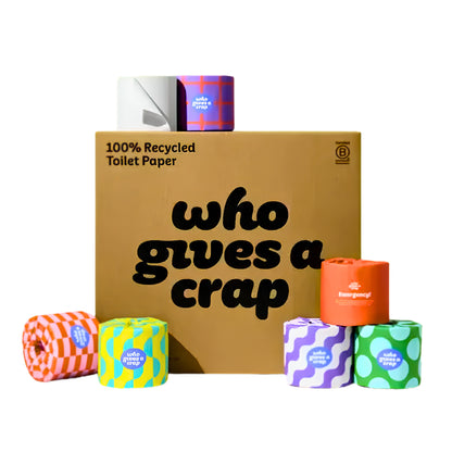 A box of toilet paper rolls on a white background, with the front text reading 'who gives a crap' and '100% Recycled Toilet Paper' on the side. Several colorful toilet paper rolls are arranged around the box, including orange, yellow, purple, green, and orange plaid designs.