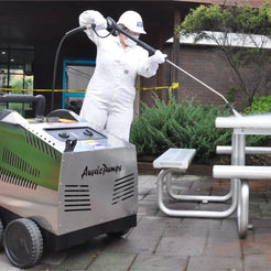 A person in a white protective suit and hard hat operates a pressure washer machine outdoors, spraying a stainless steel picnic table. The machine has 'Aussie Dumps' printed on the side, and the background shows a brick building, greenery, and yellow caution tape.