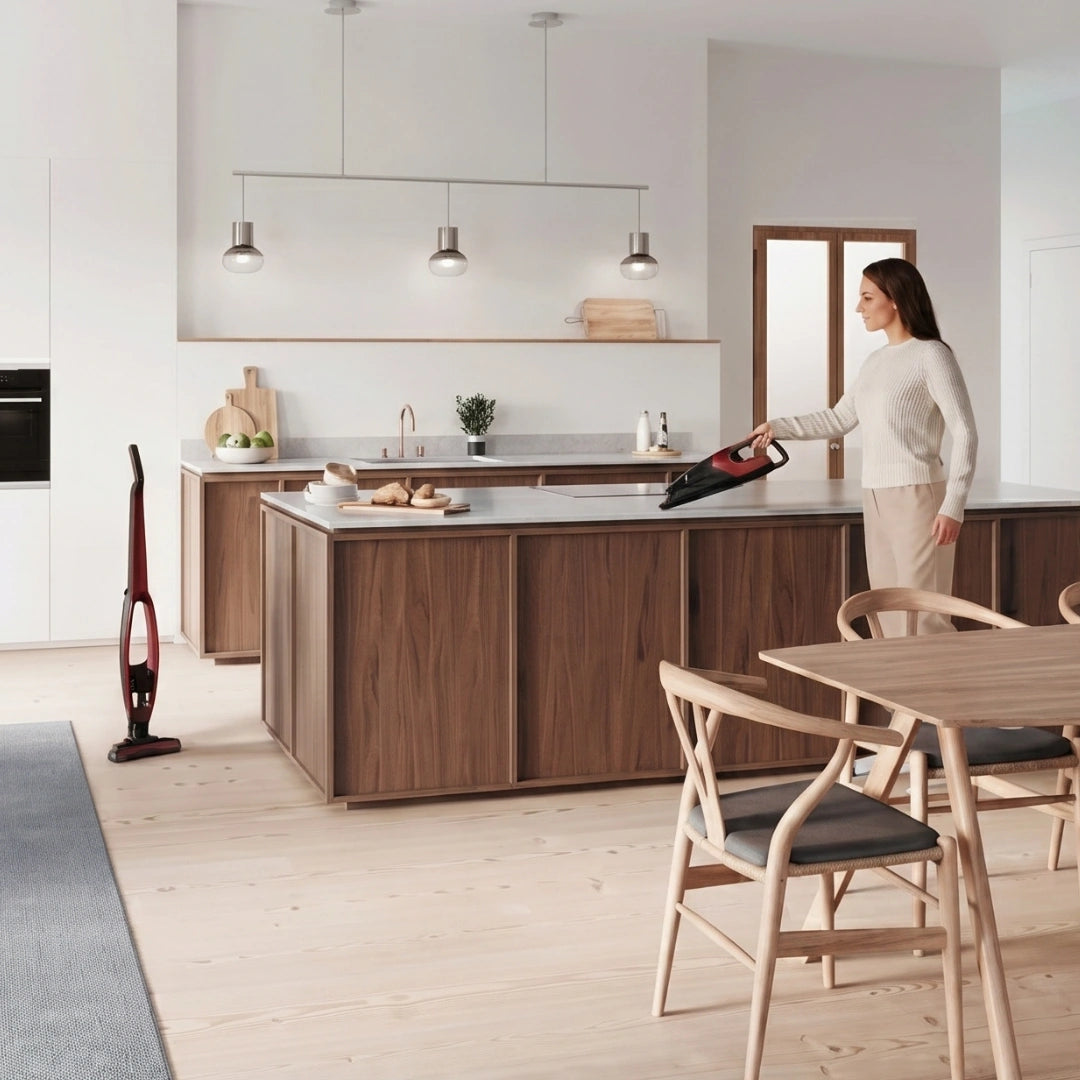 A woman in a modern kitchen uses a cordless stick vacuum cleaner, with another vacuum standing on the floor nearby. The kitchen features dark wood cabinets, a light wood island, and a dining area in the foreground.