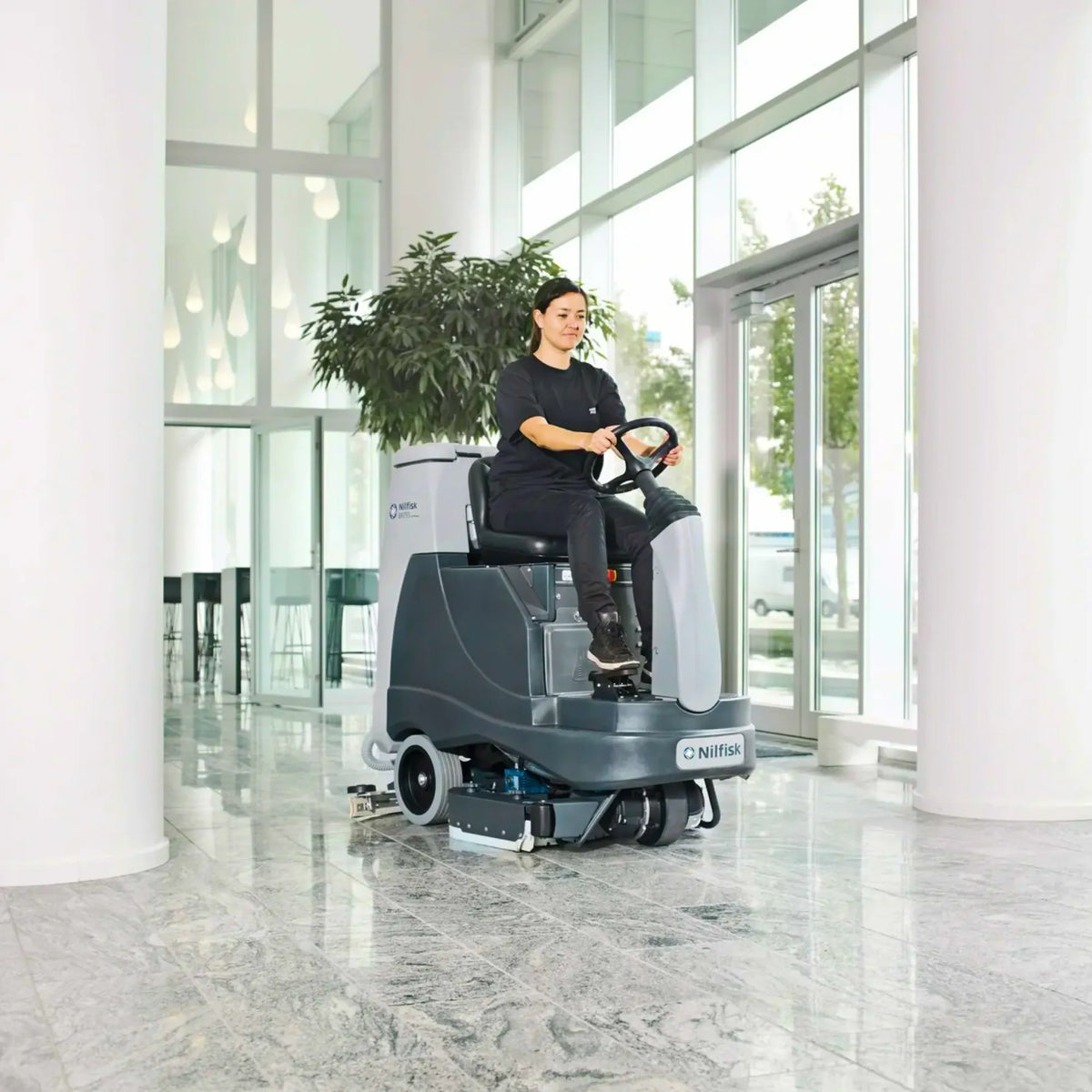 A person wearing a black uniform sits on a gray and white Nittany self-driving floor cleaner inside a modern, bright office lobby with marble floors and large windows.