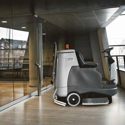 A NETAUSA AM 1100i floor care machine with a driver seat, positioned on a polished wood floor inside a modern glass-walled office space.