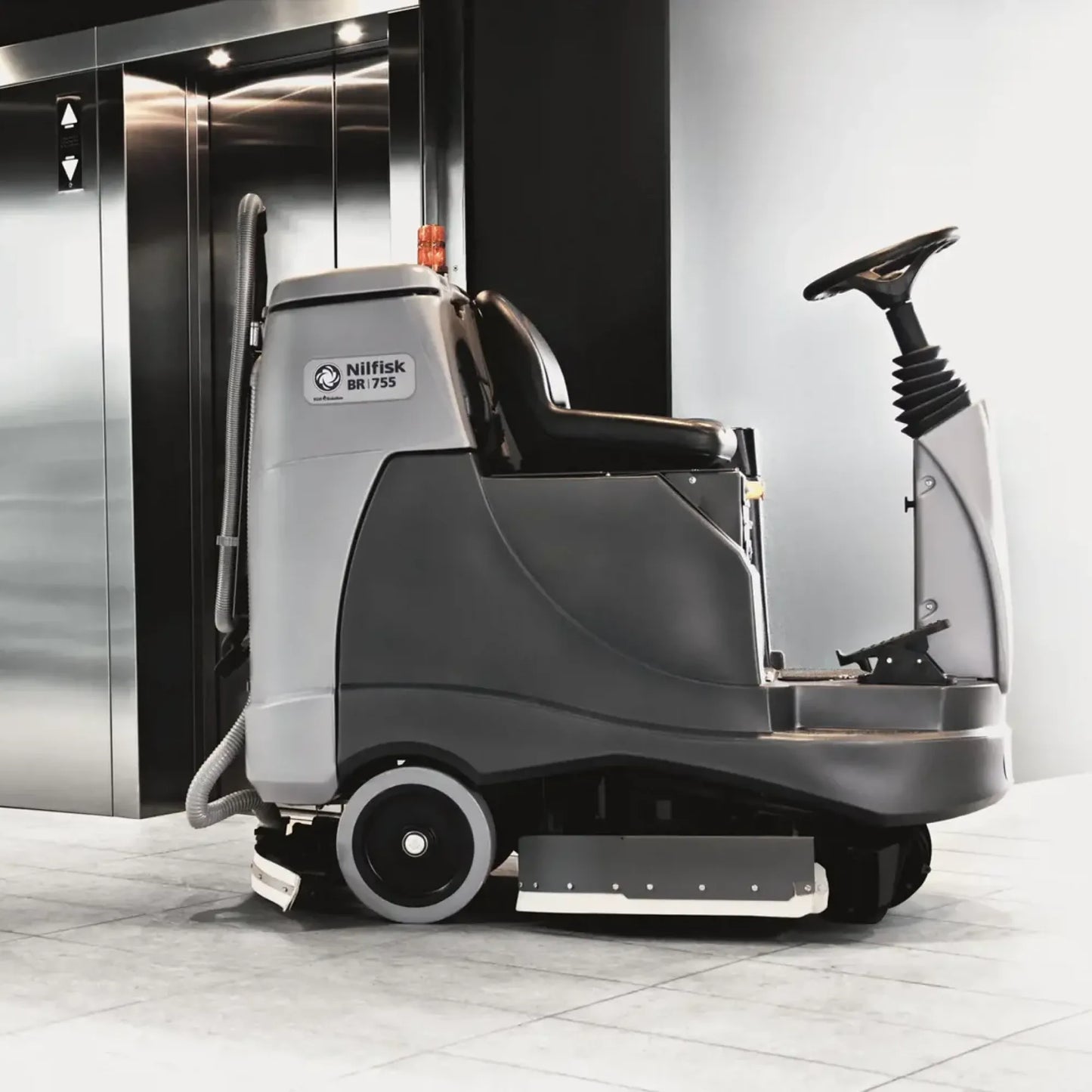 A Nilfisk WD 750 floor care vehicle with a driver seat, steering column, and front brush bar, positioned in front of an elevator lobby on a tiled floor.