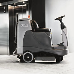 A Nilfisk WD 750 floor care vehicle with a driver seat, steering column, and front brush bar, positioned in front of an elevator lobby on a tiled floor.