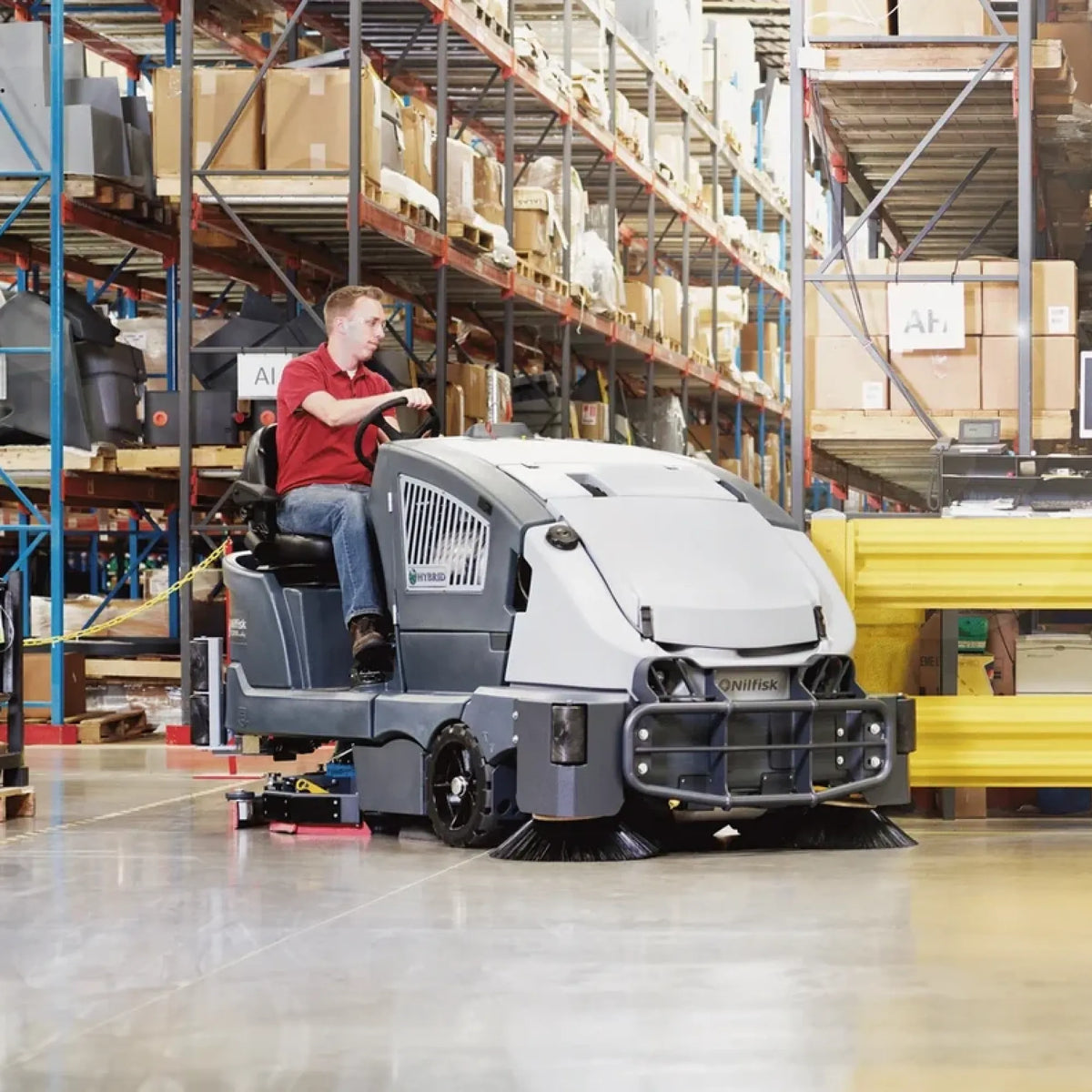 A person in a red shirt and jeans drives a compact warehouse floor cleaner robot with rotating brushes, operating it inside a warehouse aisle with tall metal shelving and cardboard boxes in the background.