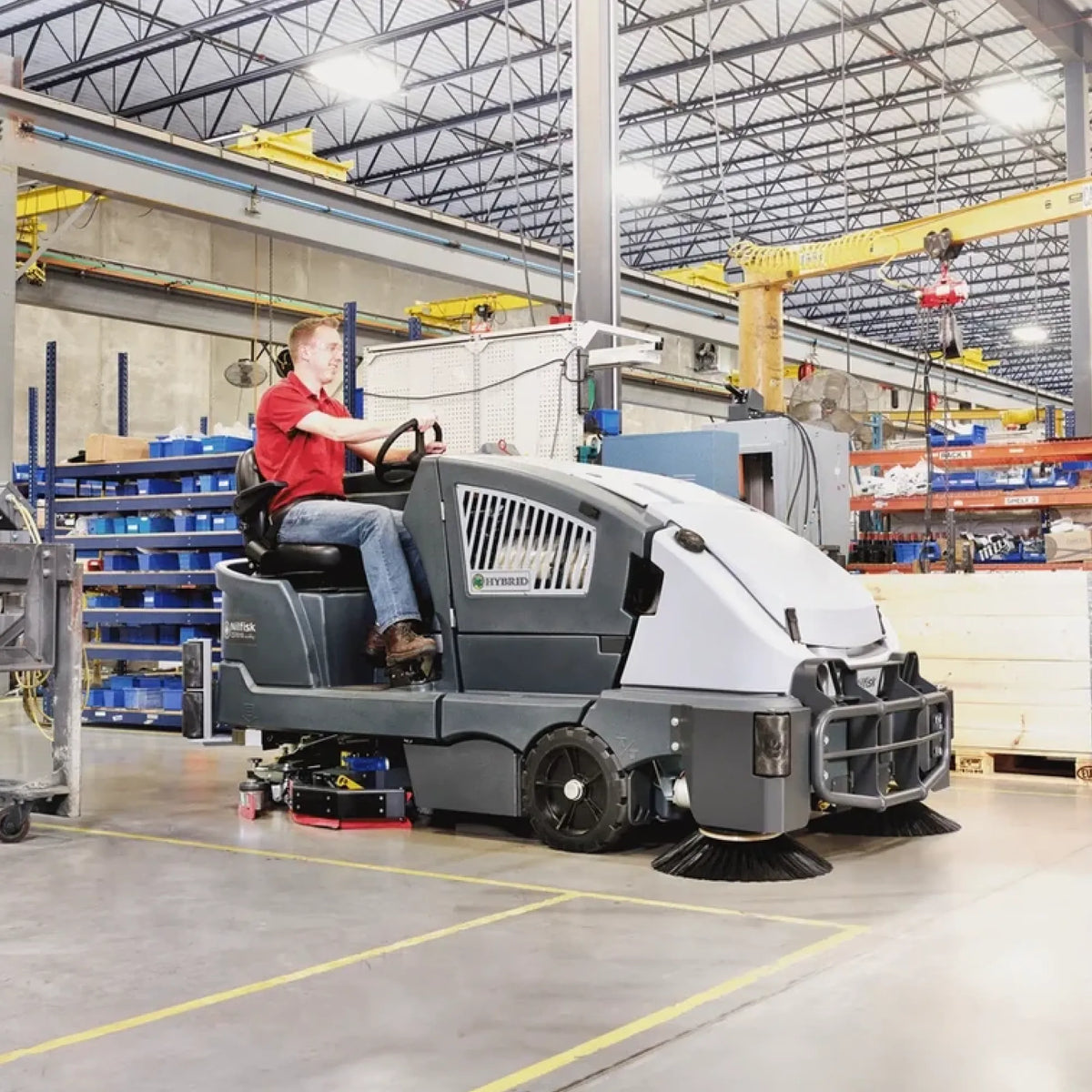 A person wearing a red shirt and jeans drives a gray and white industrial sweep truck with brush brushes on the front and side, inside a large warehouse with steel beams and storage shelves.