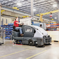 A person wearing a red shirt and jeans drives a gray and white industrial sweep truck with brush brushes on the front and side, inside a large warehouse with steel beams and storage shelves.