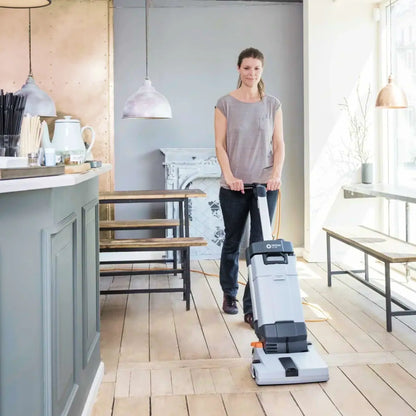 A woman pushes a white and gray floor cleaner with a long handle in a bright, modern kitchen or dining area with light wood floors.
