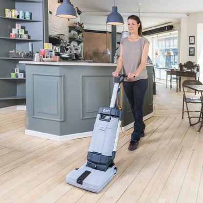 A woman pushes a large upright floor cleaner with a long handle in a bright, modern shop or cafe interior with light wood floors and a gray counter.