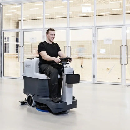 A man wearing a black t-shirt and dark pants is driving a compact floor-washing machine inside a brightly lit indoor sports hall with glass doors and volleyball nets in the background.