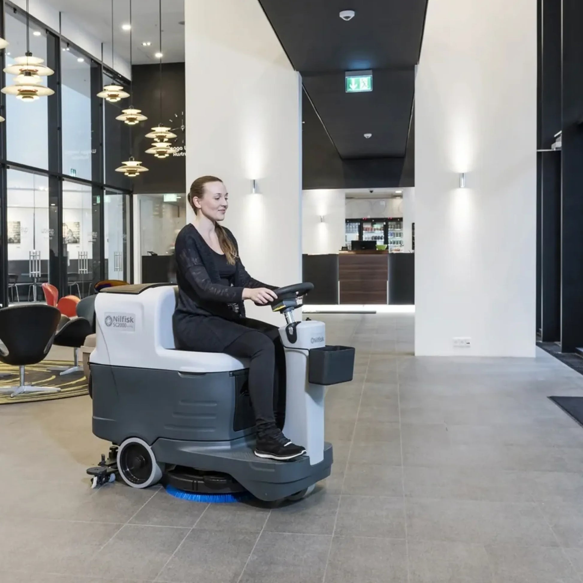 A woman wearing a black outfit sits on a white and gray Oeffag drive-in floor scrubber in a modern office lobby with gray tiled floors and black-and-white walls.