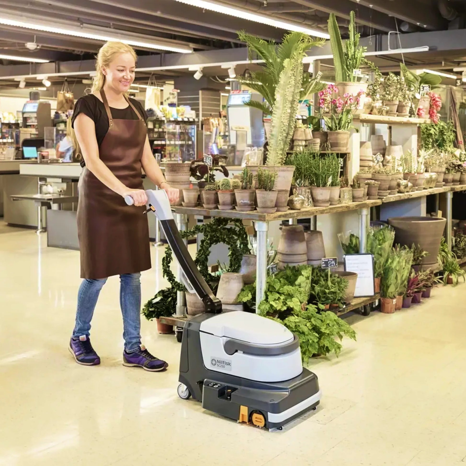 A woman wearing a brown apron and jeans operates a compact floor scrubber in a bright retail store aisle filled with potted plants.